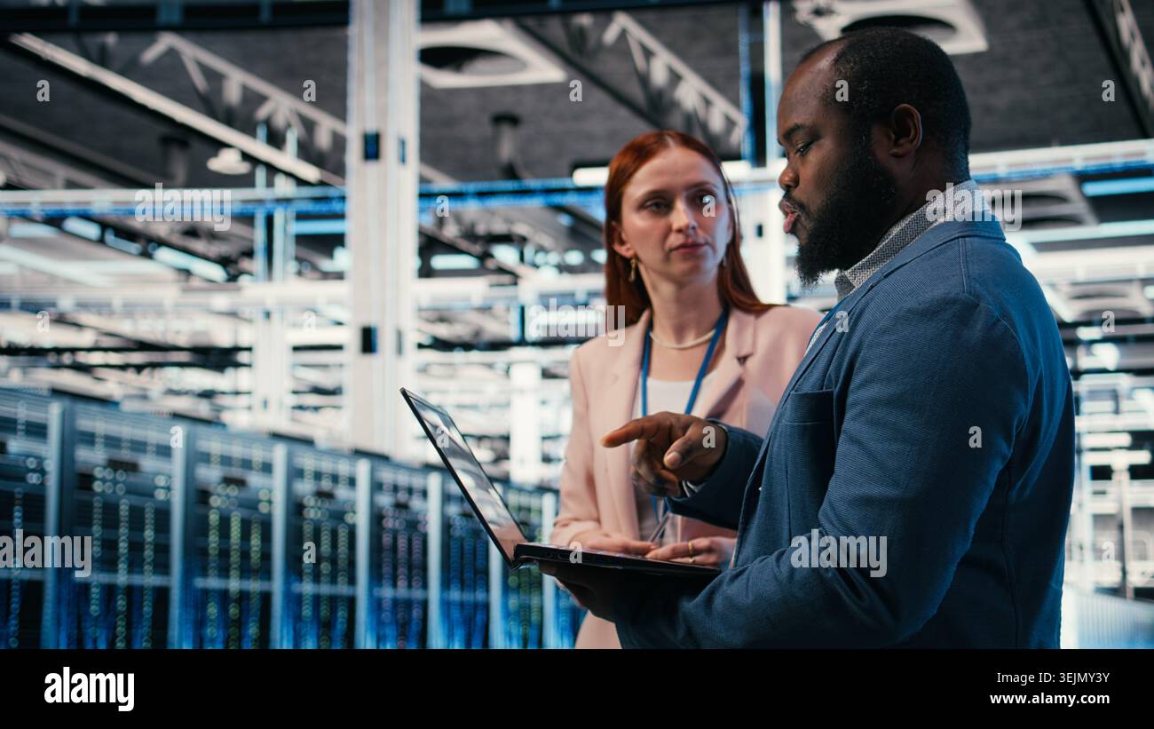 Engineering team solving tasks in server farm using laptop, doing network management virtualization. Teamworking data center colleagues oversee IT infrastructure supercomputers with notebook, camera A Stock Photo