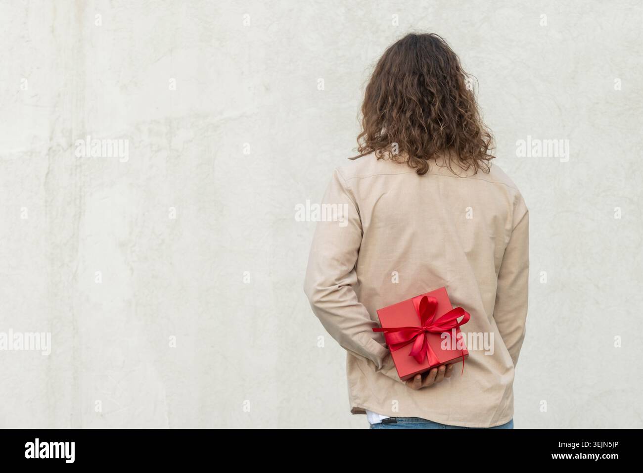 Man hiding red gift box behind his back against white wall Stock Photo