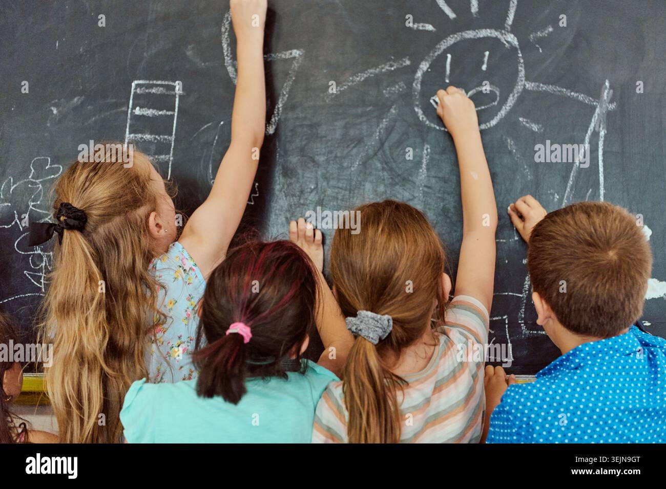 Four elementary students draw with chalk on a classroom blackboard, collaborating and expressing creativity while learning through play. Back-view cap Stock Photo