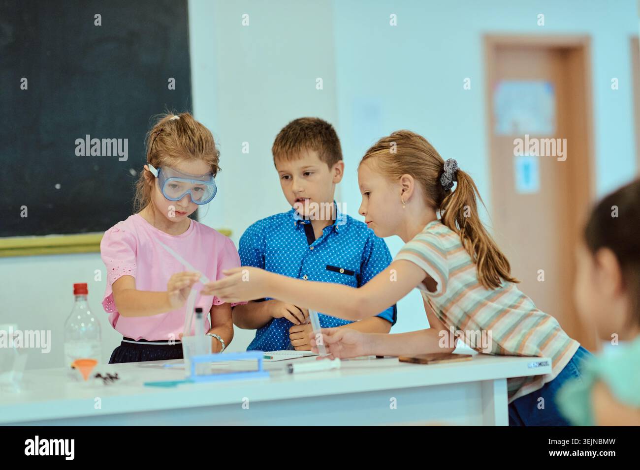 Three elementary students collaborate on a hands-on science experiment in a bright classroom, using goggles and simple lab equipment while practicing Stock Photo