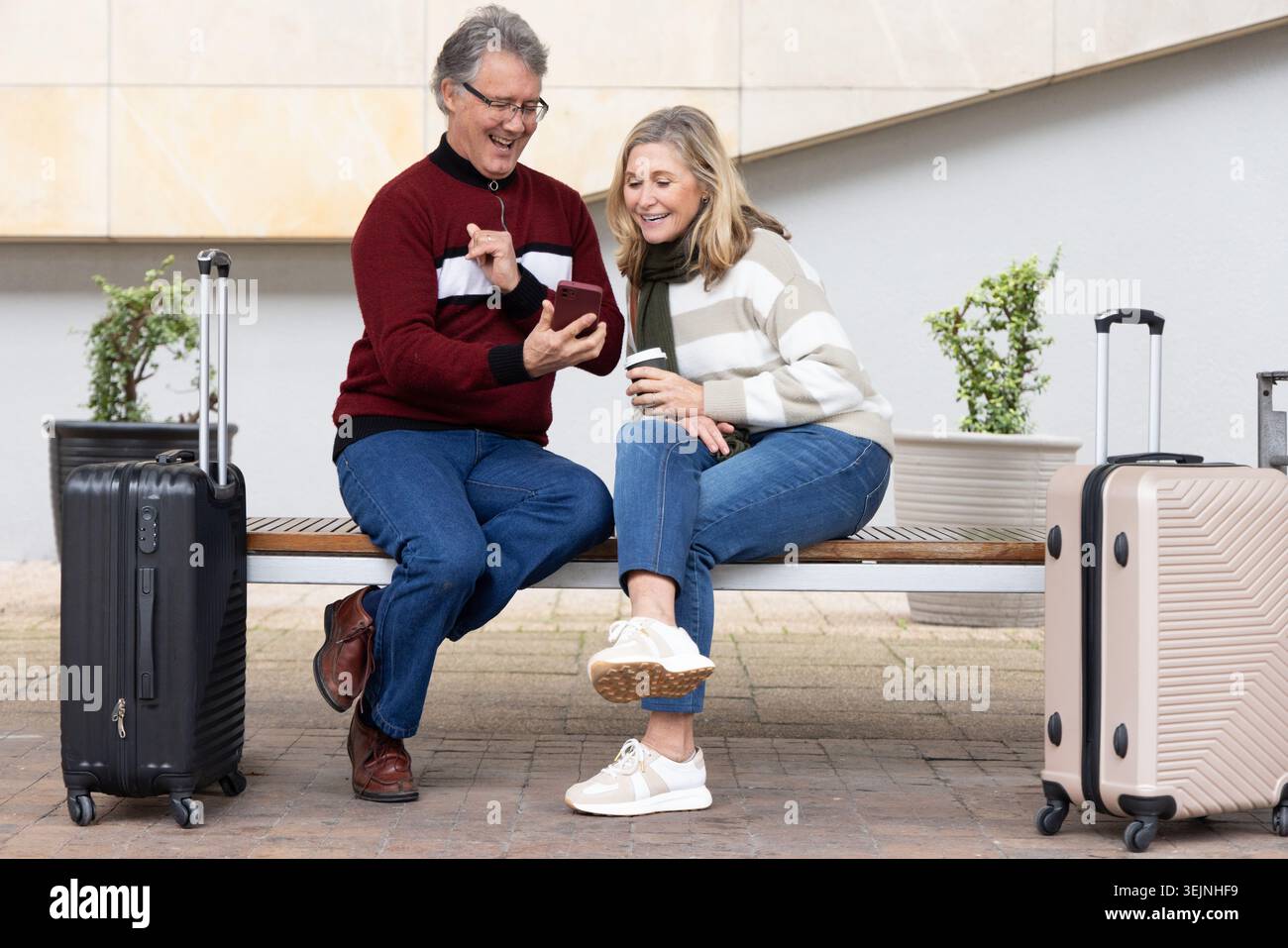 Senior couple sitting on bench at station, holding smartphone and coffee, black and beige suitcases. Retiree, duo, luggage, phon Stock Photo
