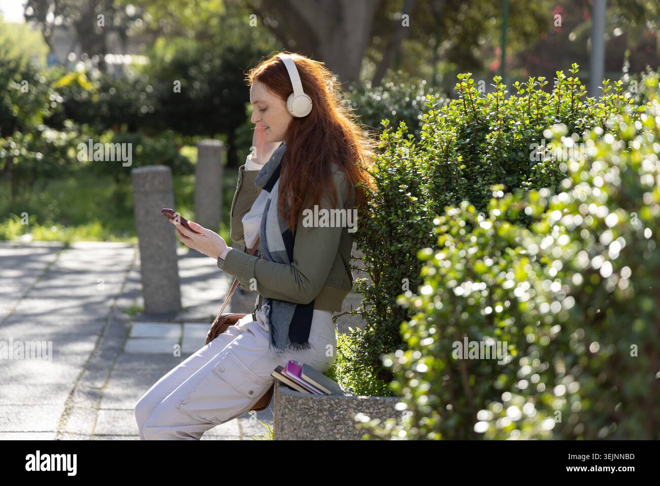 Adult female sitting on planter edge in park, listening on headphones, using phone, books and bag. Smartphone, earphones, notebook, bench, urban, natu Stock Photo