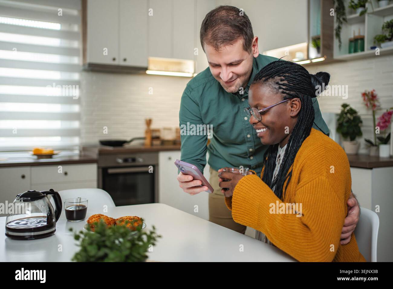 Diverse couple smiling, using smartphone, having breakfast in kitchen Stock Photo