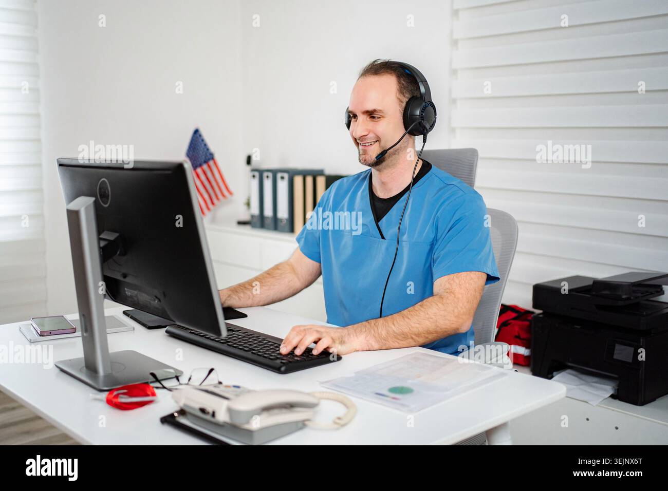 Man in scrubs smiling, using a computer and headset for remote medical work Stock Photo
