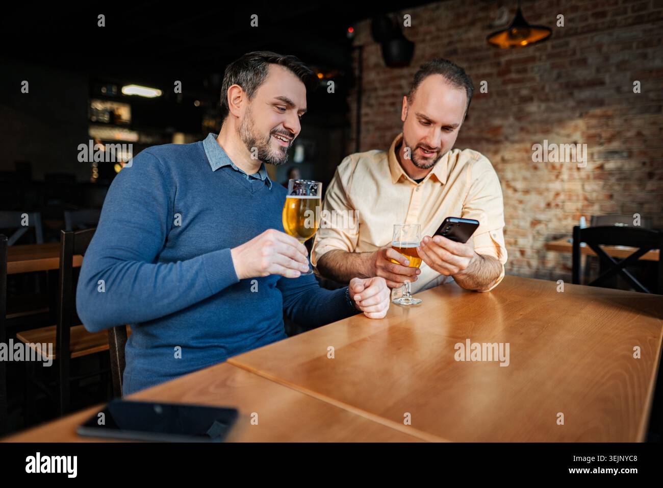 Two friends relaxing at a bar, drinking beer, using smartphone Stock Photo