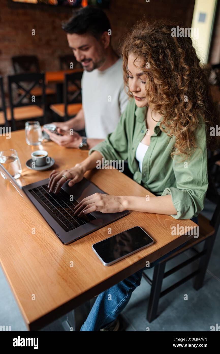 Young woman working on laptop in cafe while man using smartphone Stock Photo