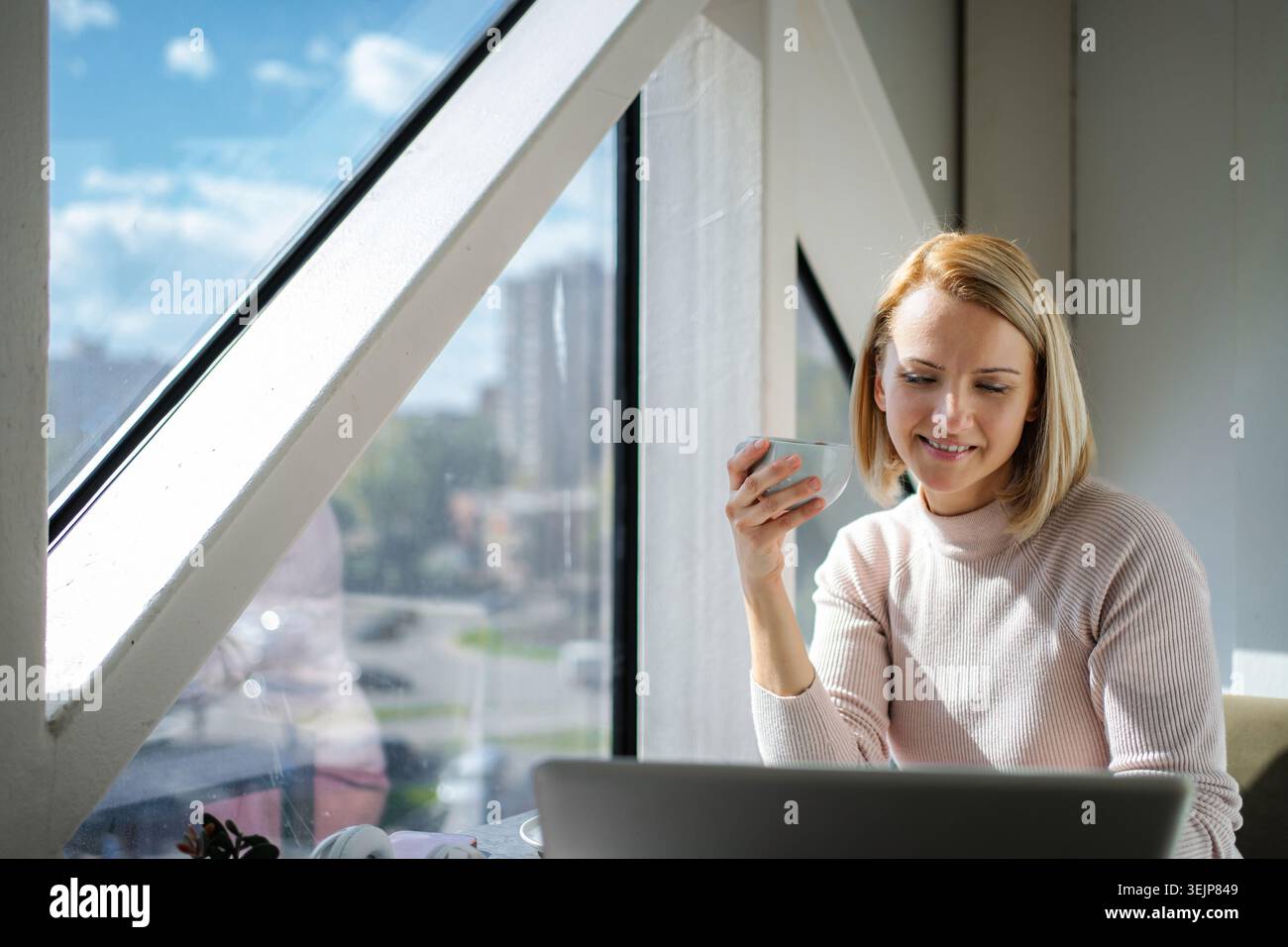 Woman drinking coffee while working on laptop by window Stock Photo