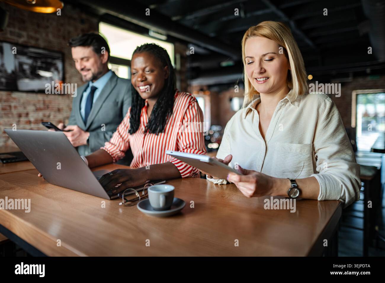 Three diverse business professionals working, communicating using digital devices in a modern cafe Stock Photo