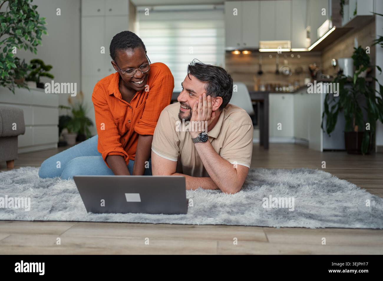 Happy multi-ethnic couple lying on carpet using laptop at home Stock Photo