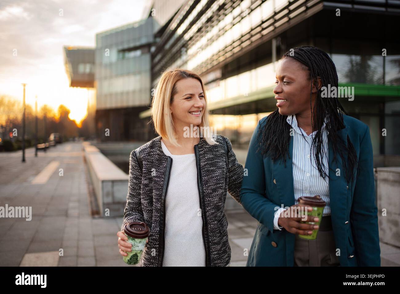 Two businesswomen walking together outdoors while having a joyful conversation and drinking coffee Stock Photo