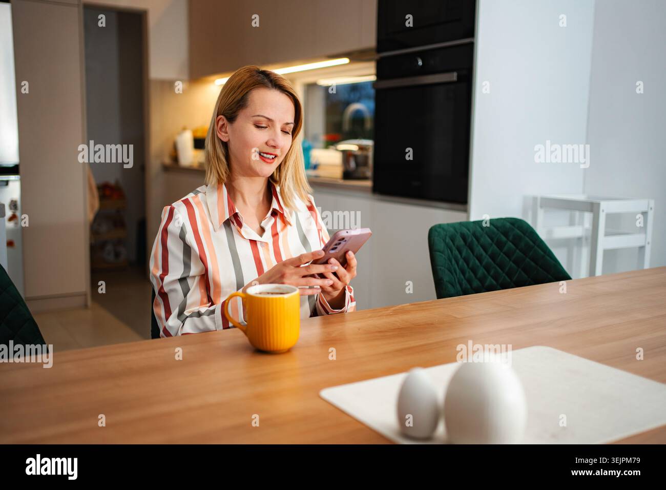 Woman drinking coffee and using smartphone in kitchen Stock Photo