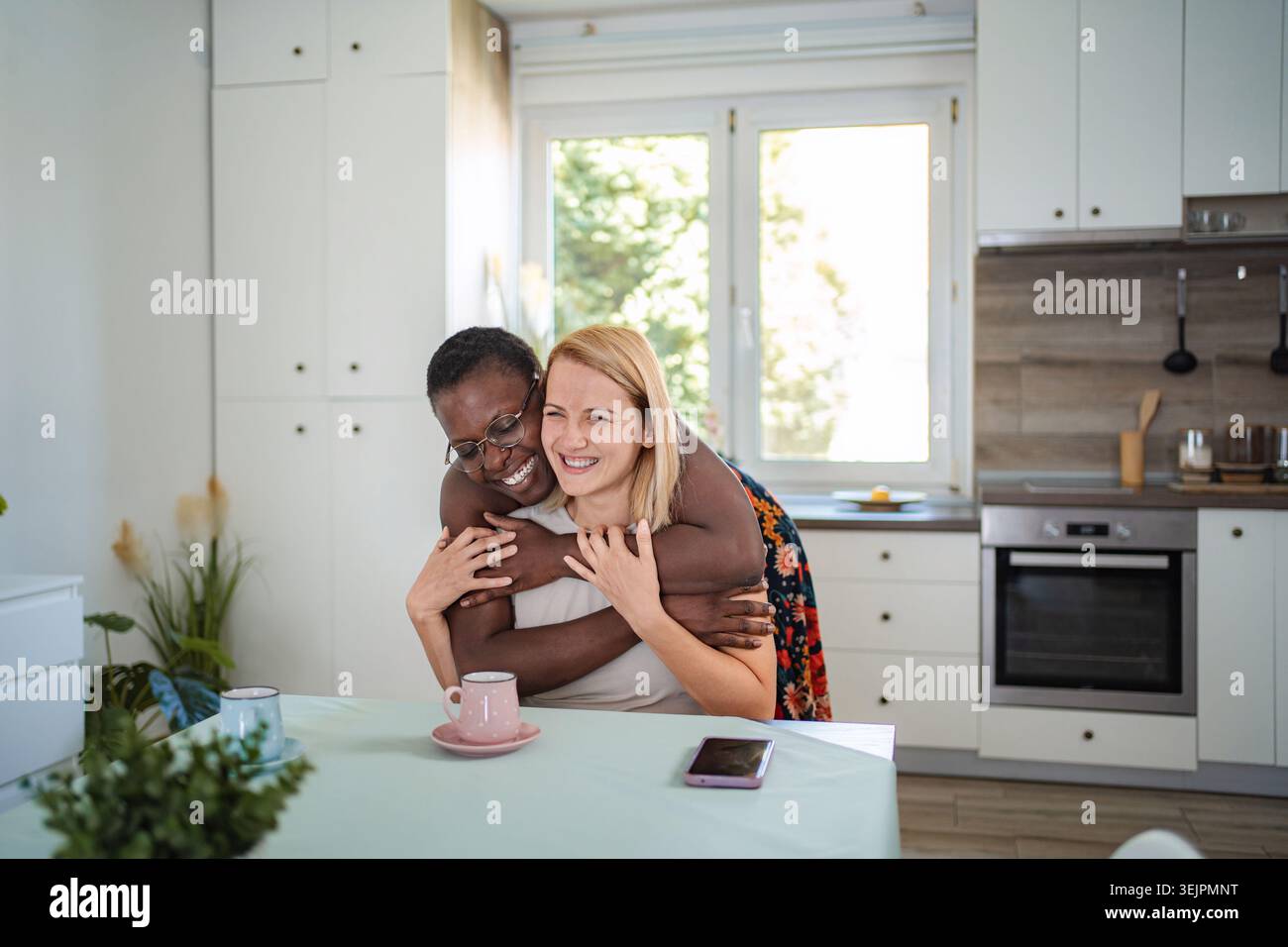 Two cheerful female friends are hugging in the kitchen Stock Photo