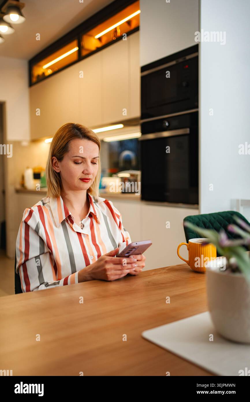 Woman using smartphone while relaxing in modern kitchen Stock Photo