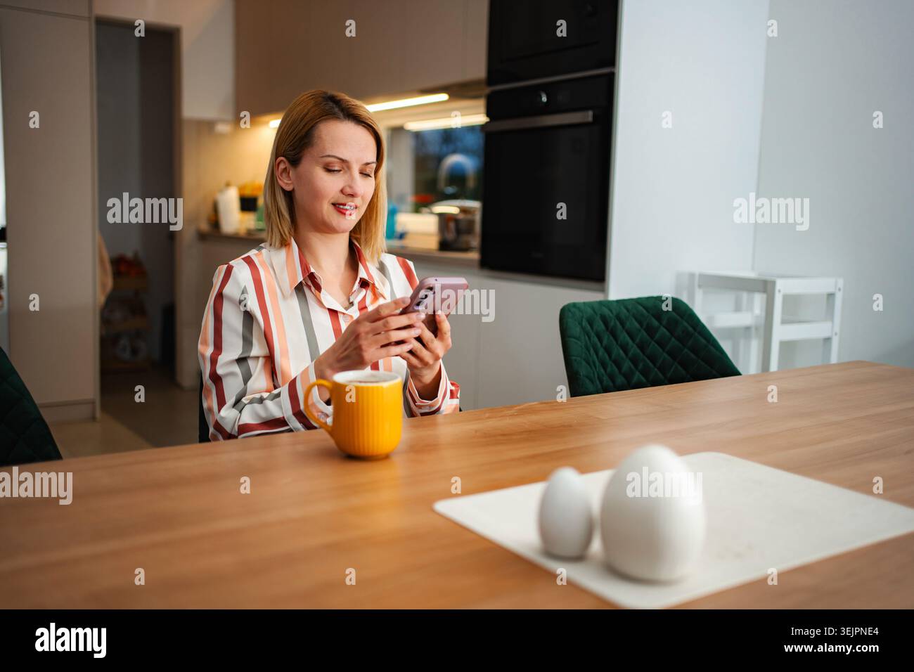 Woman using smartphone and drinking coffee in kitchen Stock Photo