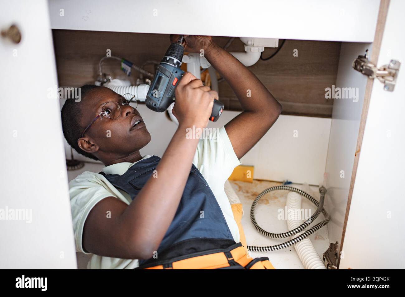Young female plumber lying down and using electric screwdriver beneath kitchen sink Stock Photo