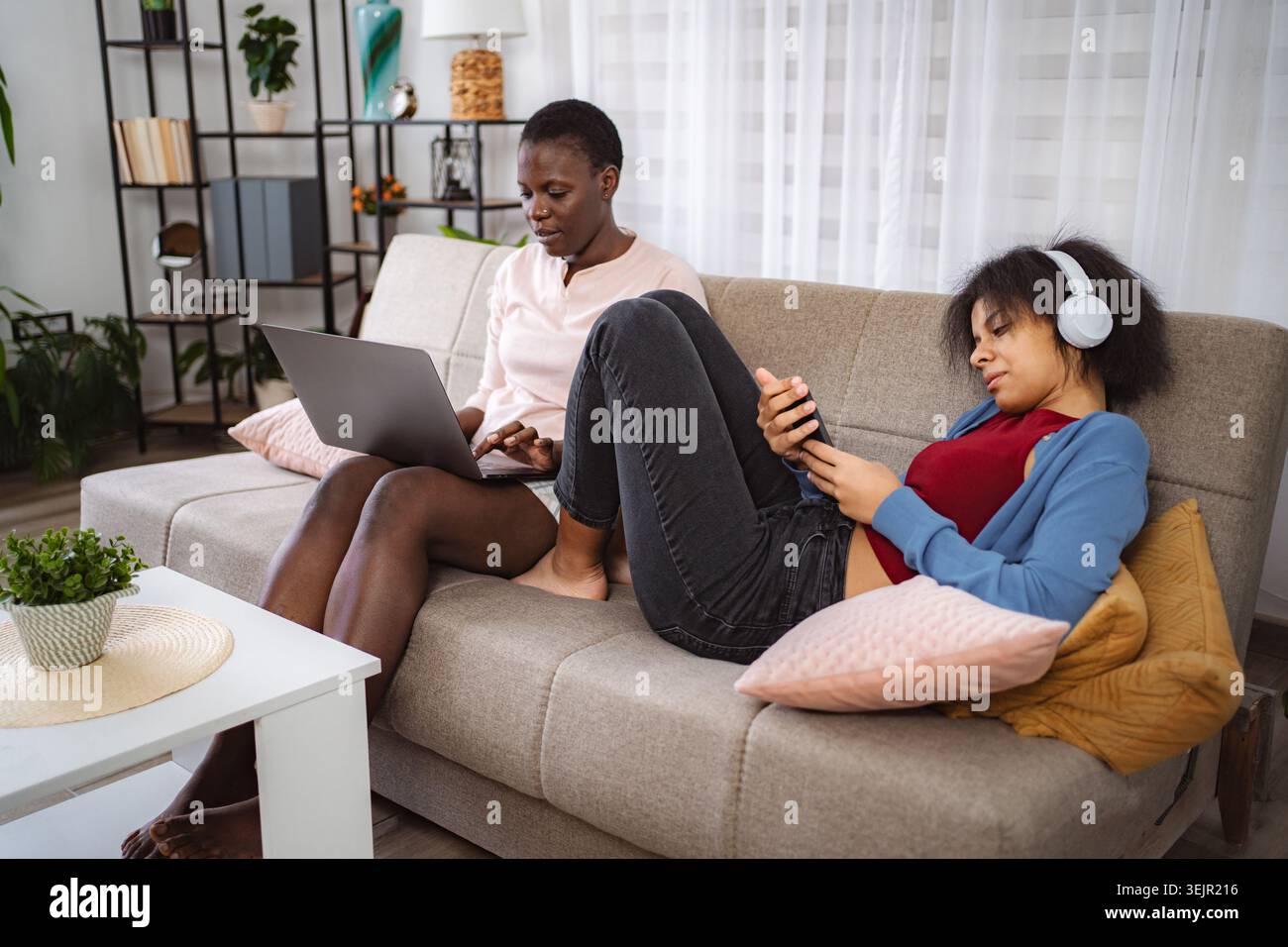 Two Young Women Relaxing on Sofa, Using Laptop and Smartphone Stock Photo