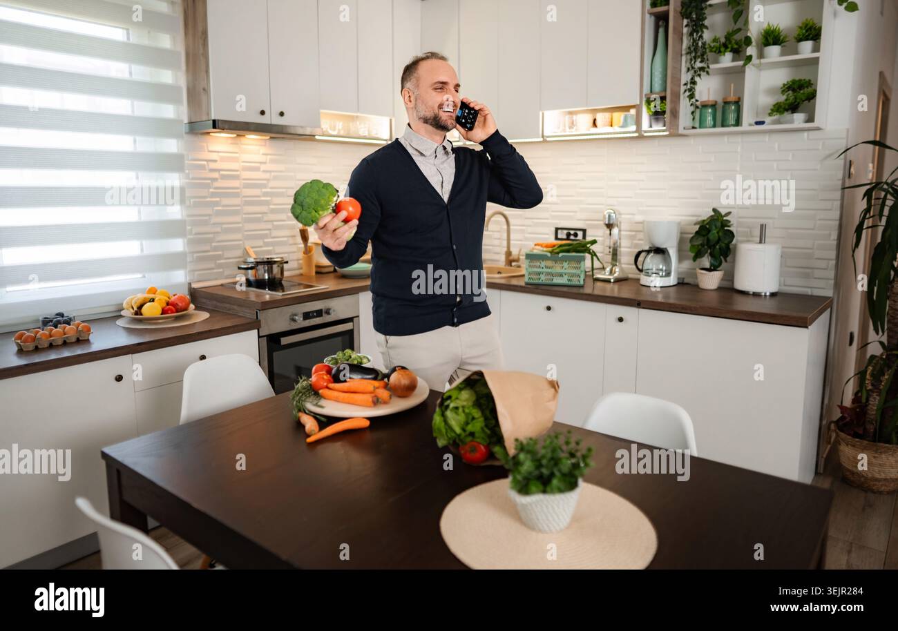 Man talking on phone holding vegetables in kitchen Stock Photo