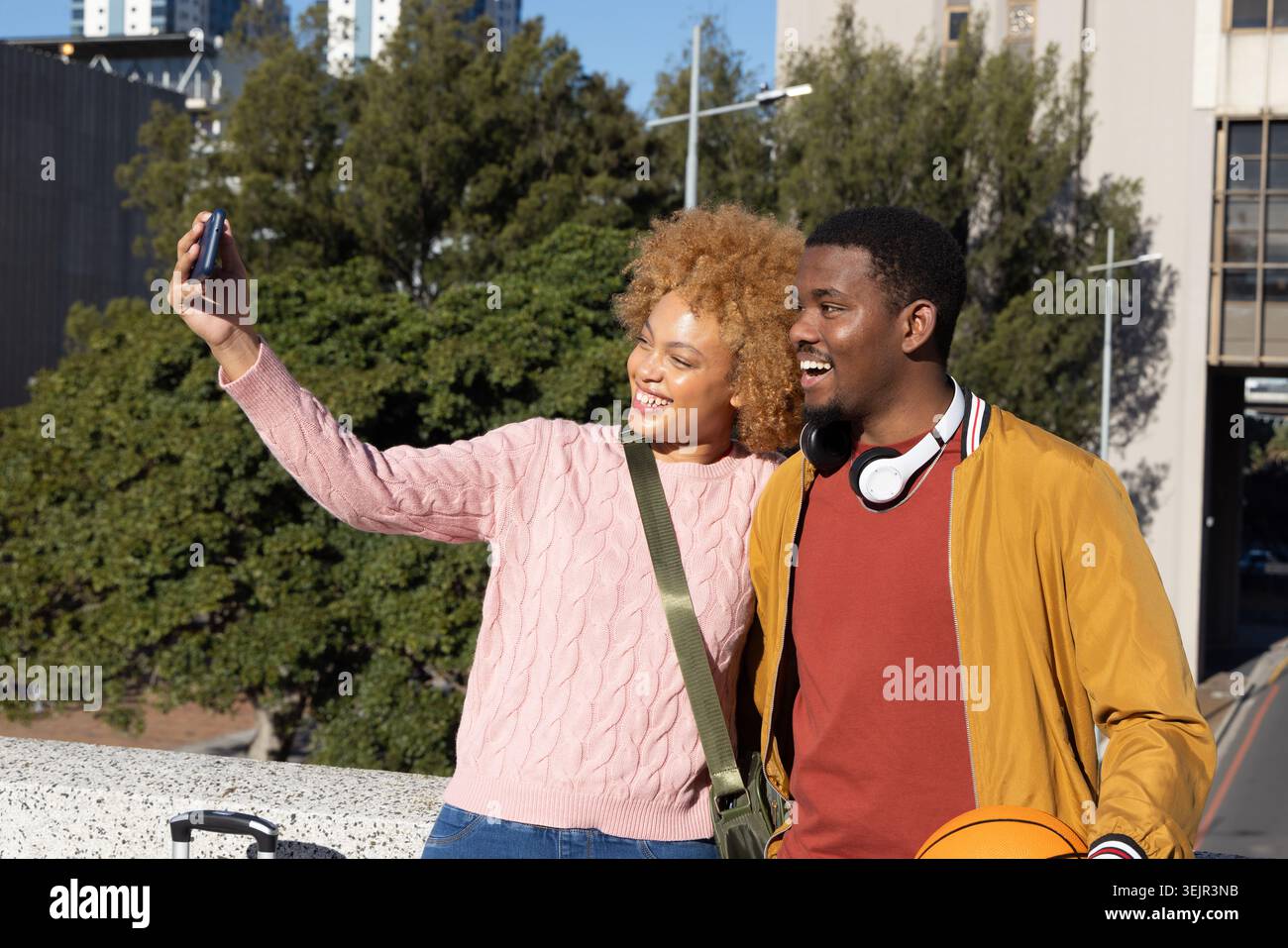 African American couple taking smartphone selfie at overlook with suitcase, man holding basketball. Duo, lifestyle, outdoor, urb Stock Photo