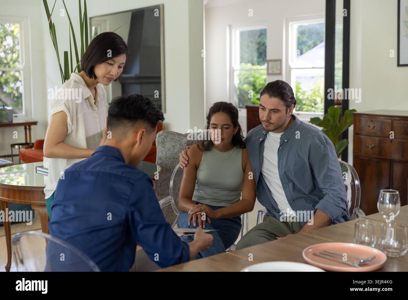 Diverse couple consulting two advisors, reviewing paperwork on wooden dining table with pen. Home, daylight, interior, greenery, Stock Photo