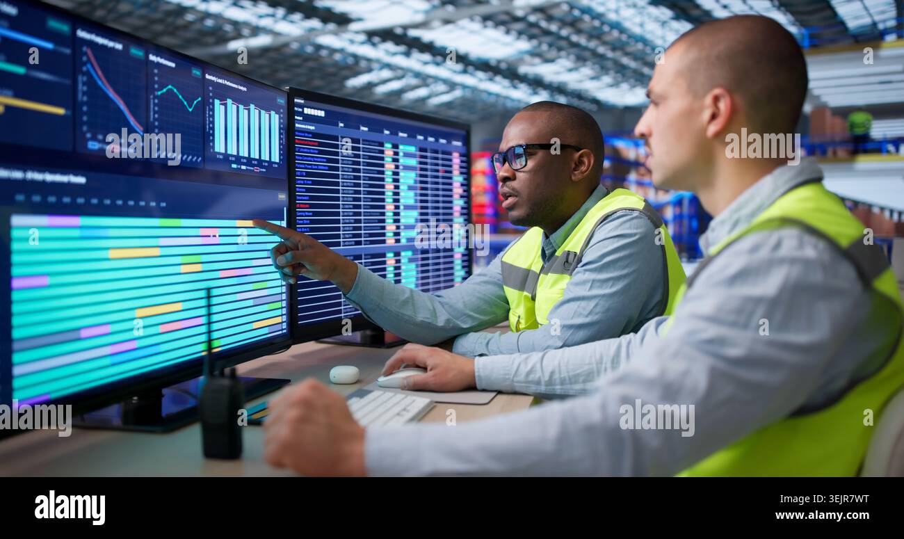 Two Workers Sit In Warehouse Managing Logistics Orders On Computers. Stock Photo