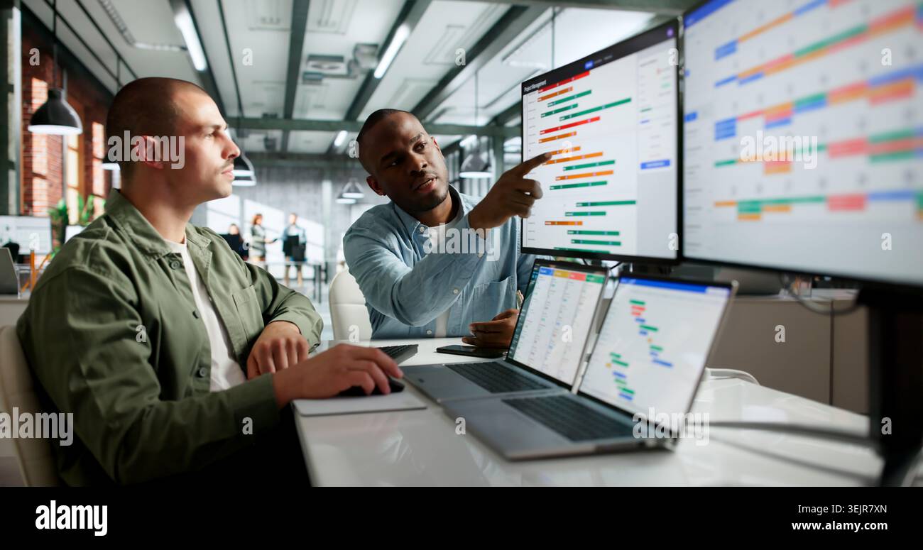 Two Team Members Discuss Project Schedule And Plan Tasks With Laptop Open. Stock Photo