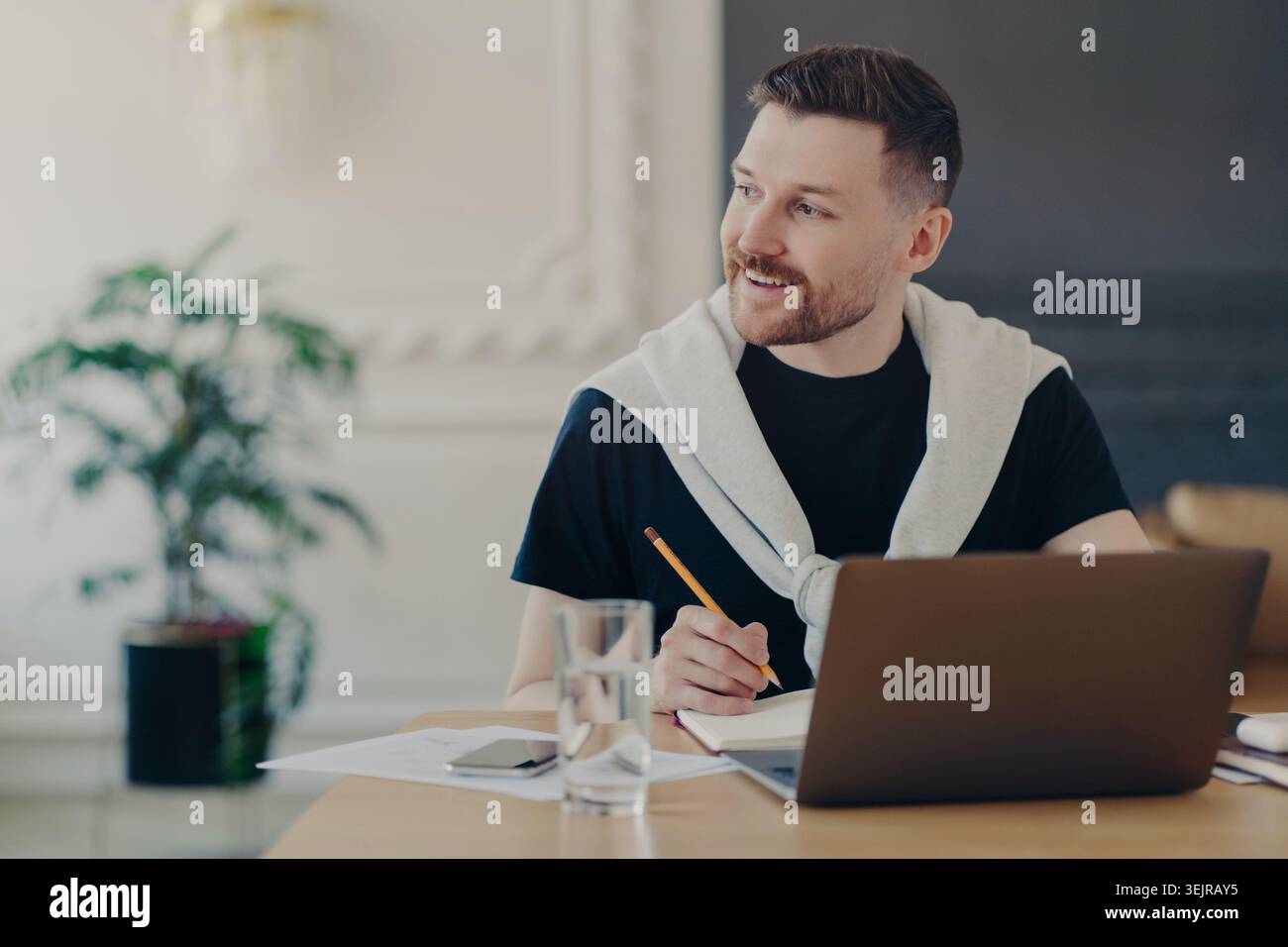 Young man writing notes while using laptop at home office desk Stock Photo