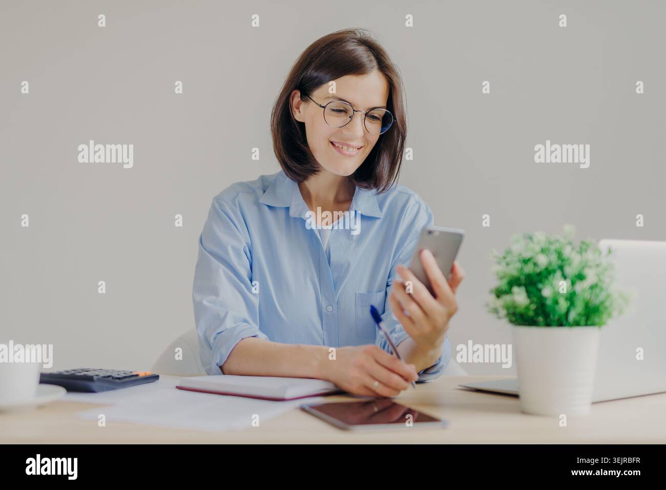 Woman Using Smartphone at Desk While Writing Notes in Bright Modern Home Office Stock Photo