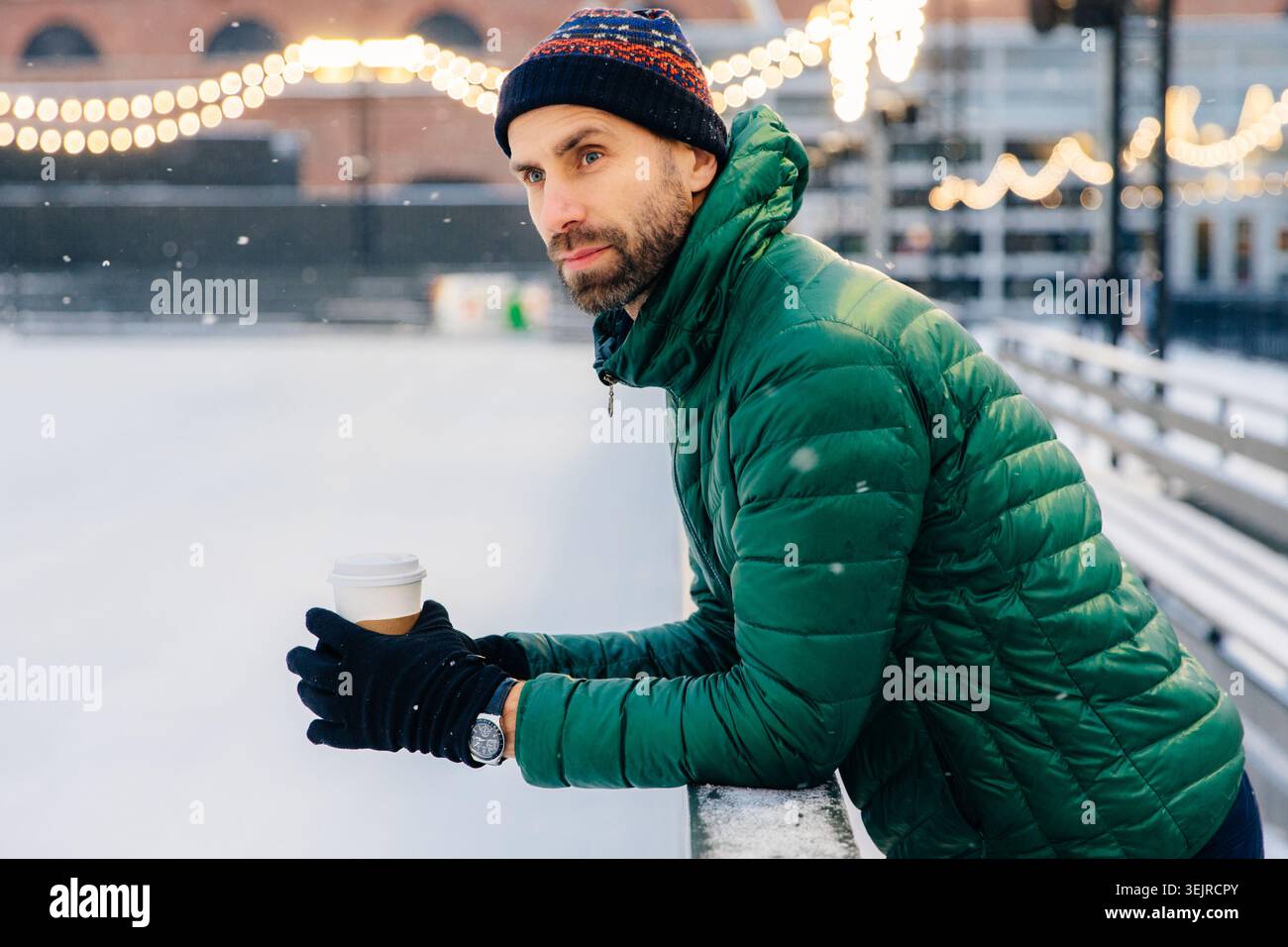 Young Man Holding Takeaway Coffee at Outdoor Ice Rink in Winter Wearing Green Jacket and Knit Hat Stock Photo