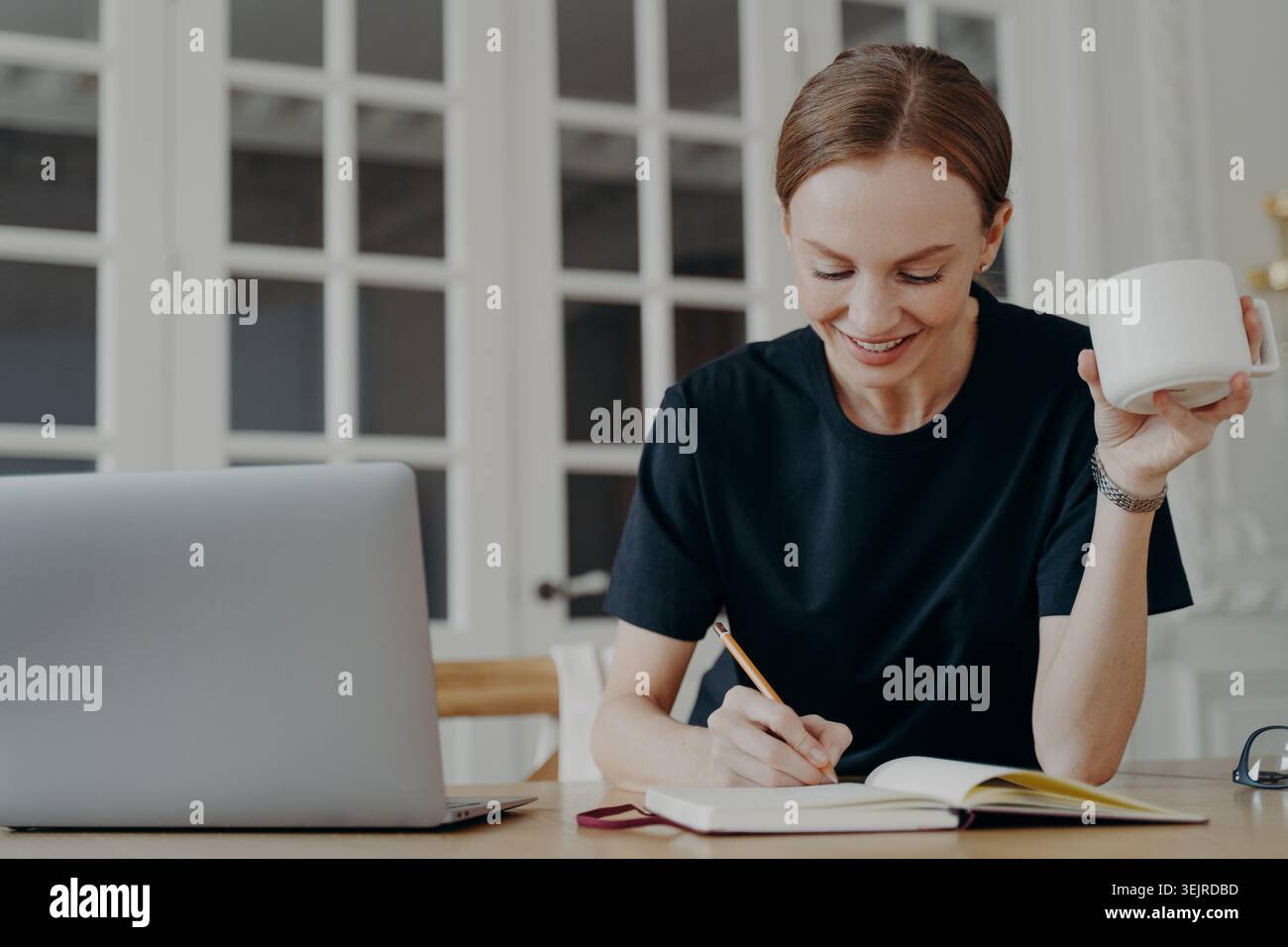 Focused Woman Taking Notes at Home Desk with Laptop and Coffee During Remote Work Stock Photo