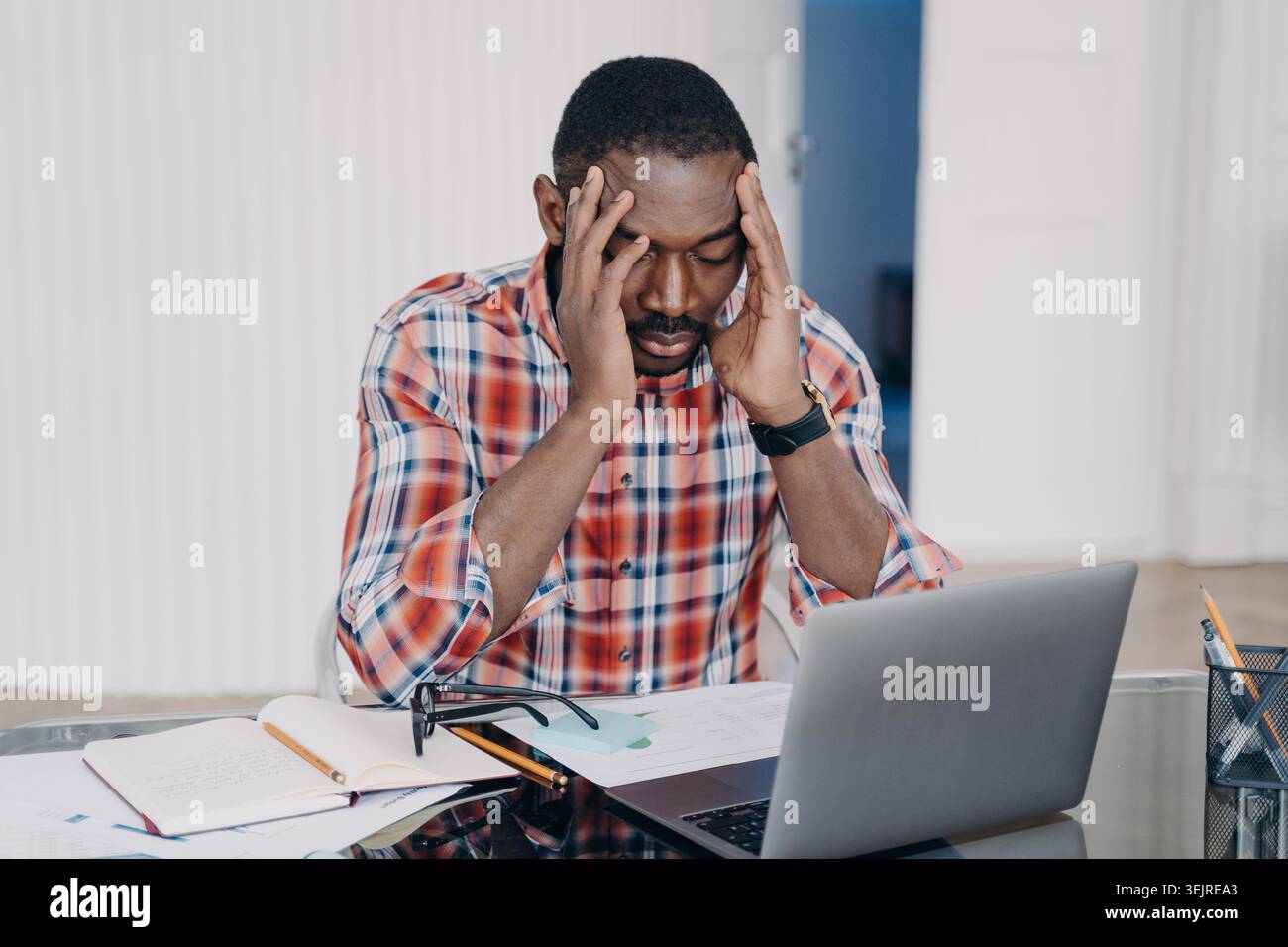 Stressed Black Man Working on Laptop at Home Office Holding Head in Hands While Feeling Overwhelmed by Papers and Notes Stock Photo