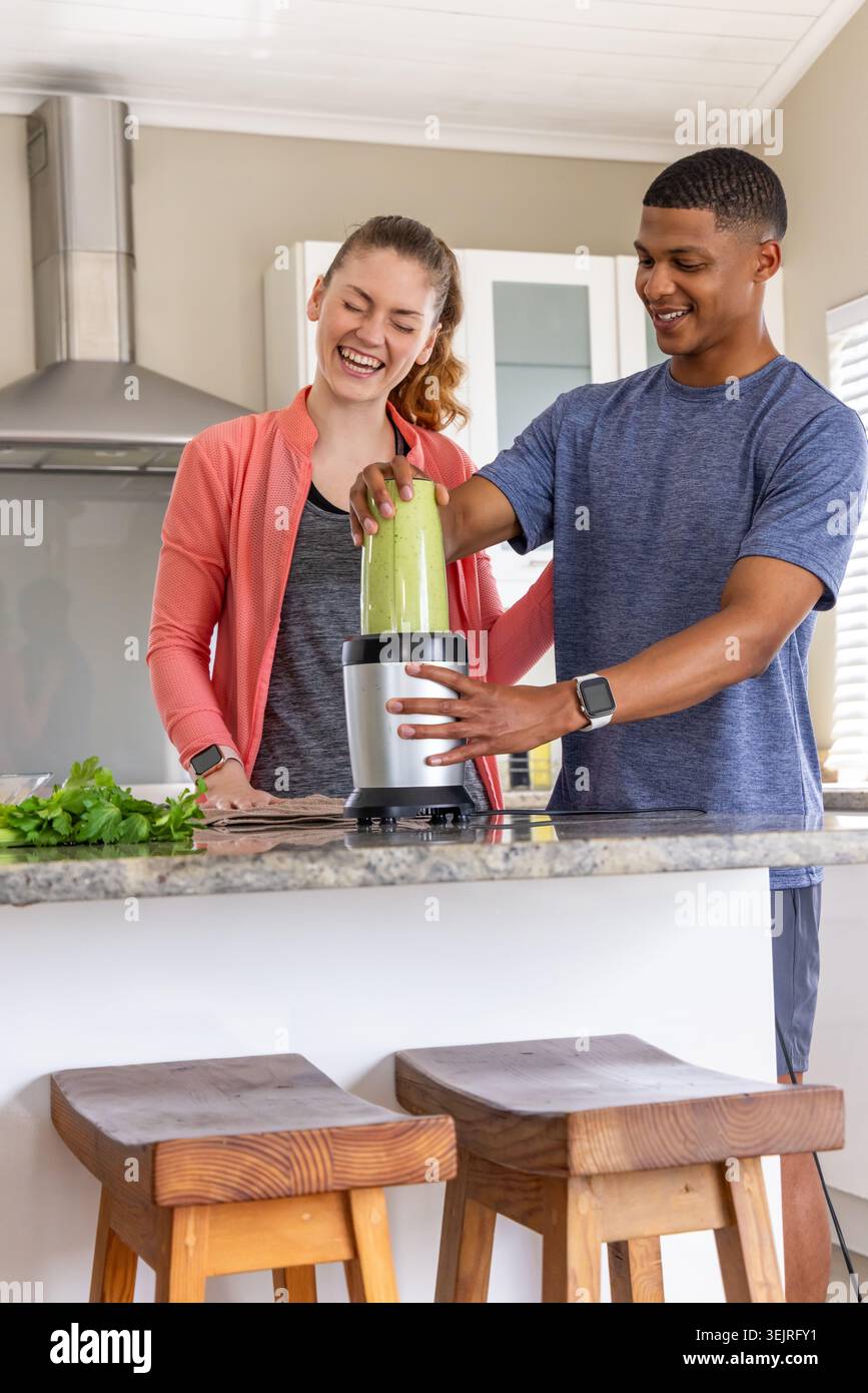 Diverse couple preparing green smoothie at kitchen island with blender, wearing coral jacket. Drink, herbs, parsley, cilantro, healthy, wellness, gran Stock Photo