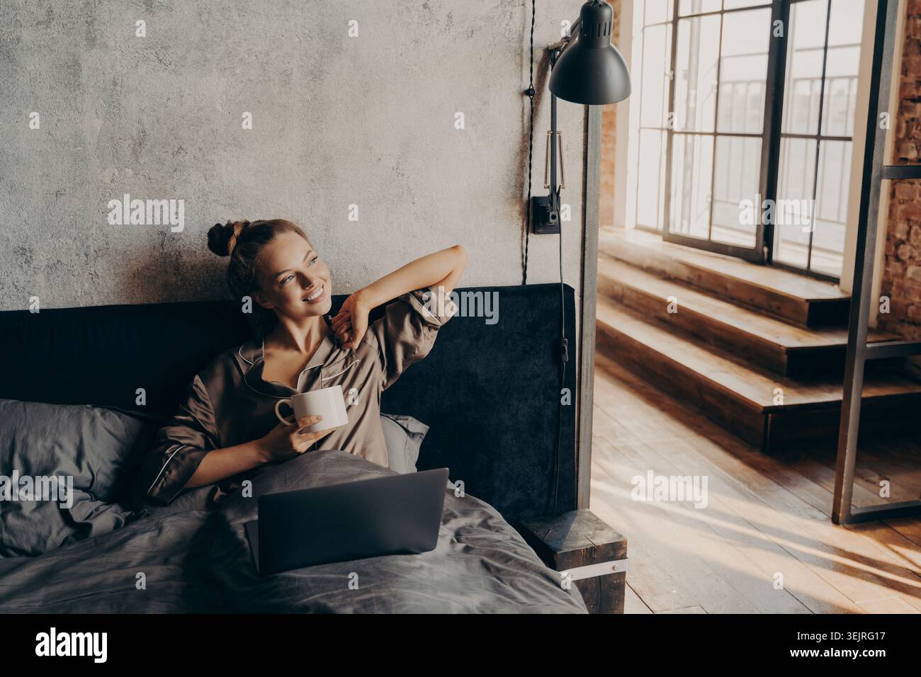 Young woman drinks coffee while using laptop in bed in loft apartment Stock Photo