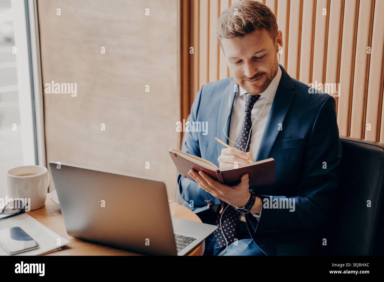 Businessman writes notes in planner beside laptop while working at cafe table Stock Photo