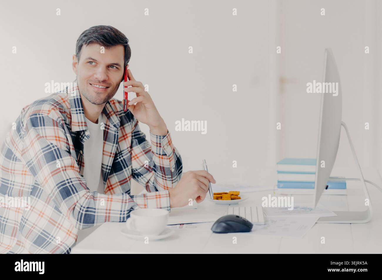 Young man talks on smartphone while working at desktop computer in bright home office Stock Photo