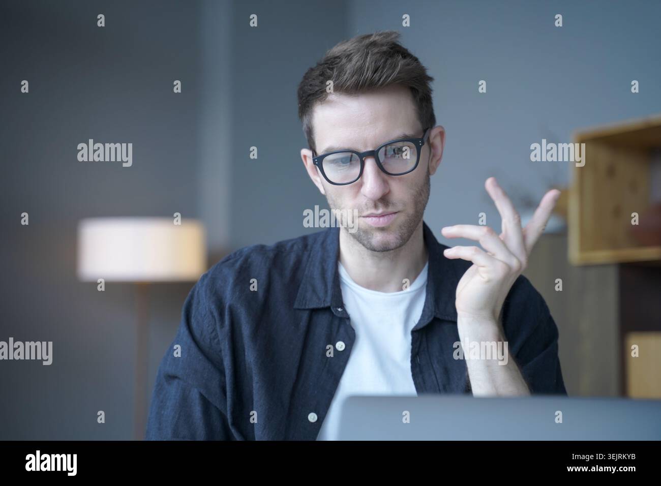 Young man with glasses gestures while using laptop in home office Stock Photo
