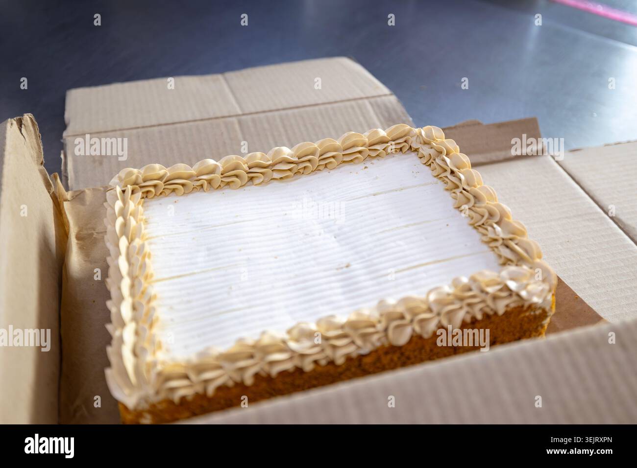 A rectangular sheet cake with white frosting and a cream-colored piped border rests inside an open cardboard box placed on a dark metal tabletop surfa Stock Photo