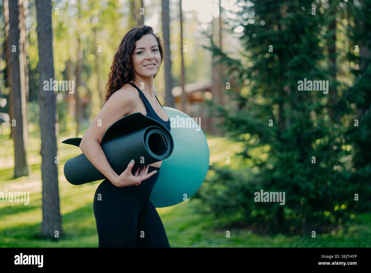 Sideways shot of beautiful curly brunette woman carries rolled up karemat and fitness ball, dressed in active wear, poses in forest or park, has fitness training outdoor, leads active lifestyle Stock Photo