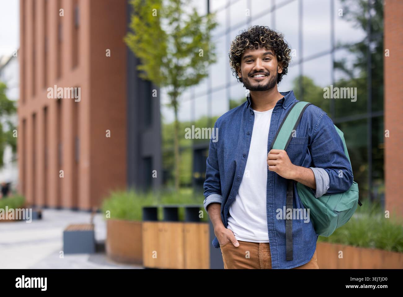 Young indian male student with curly hair smiling and looking at camera, carrying a backpack while standing outside a university building, representing education and lifestyle Stock Photo