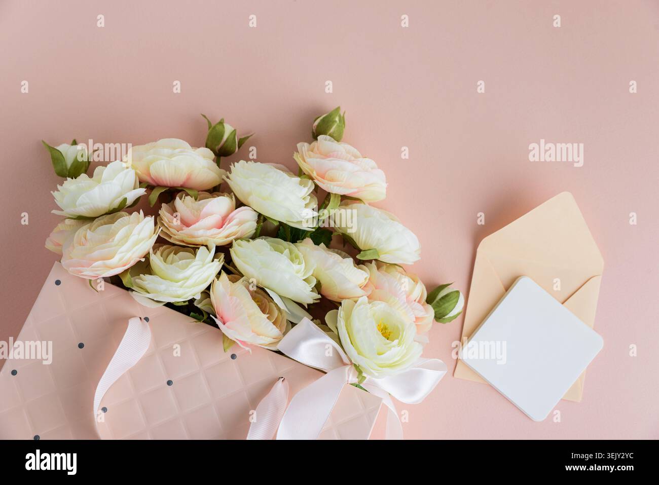 Festive composition featuring a quilted pink gift bag filled with cream and blush artificial rose flowers, accented by a white ribbon bow and gift car Stock Photo
