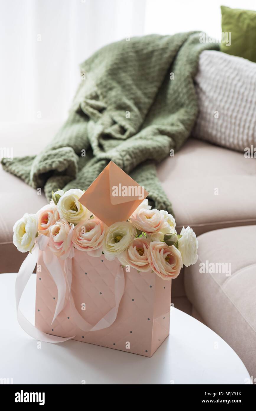 Delicate lifestyle composition featuring a quilted pink gift bag filled with cream and blush ranunculus flowers, accented by ribbon handles and decora Stock Photo