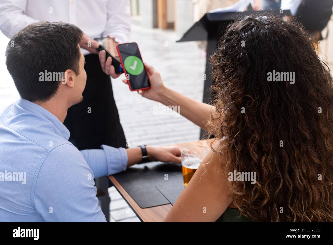 Rear view of a couple paying a restaurant bill with a smartphone using contactless technology. Waiter holds the payment terminal while the customer co Stock Photo