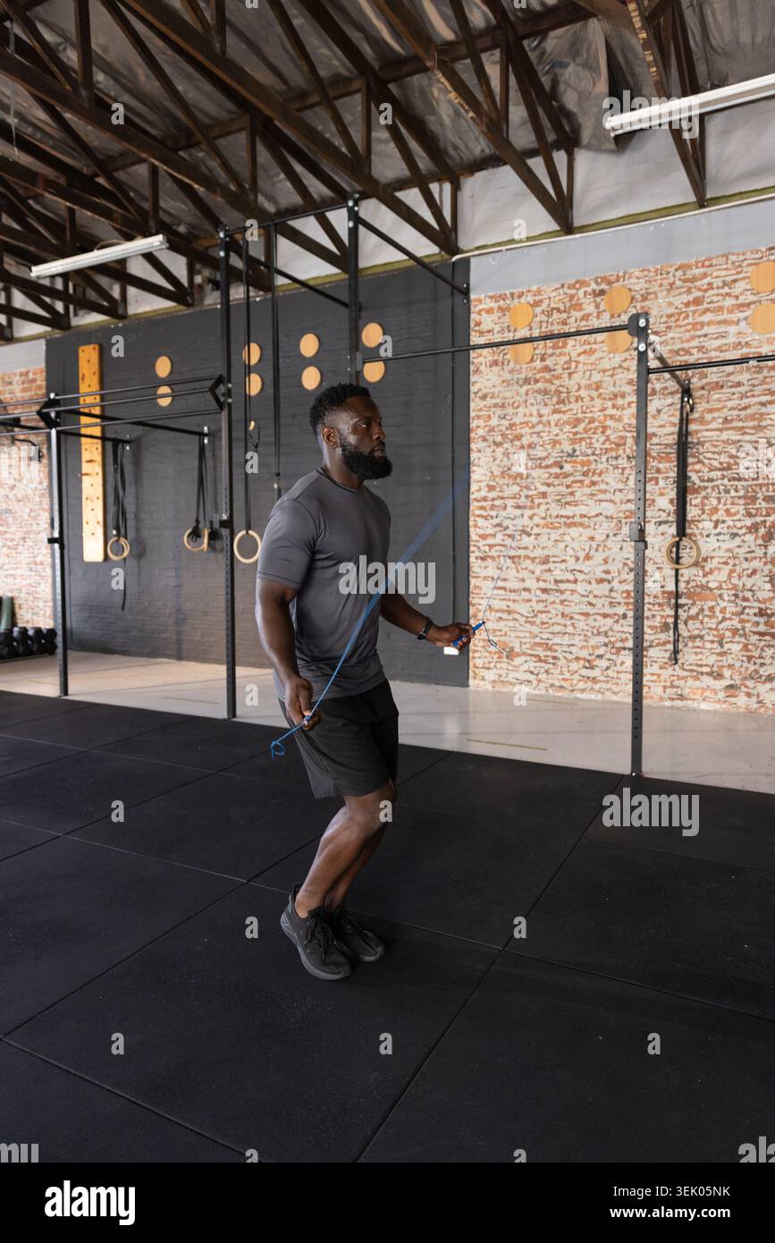 Adult African American man jumping rope with blue cord at functional gym wearing fitted gray shirt Stock Photo