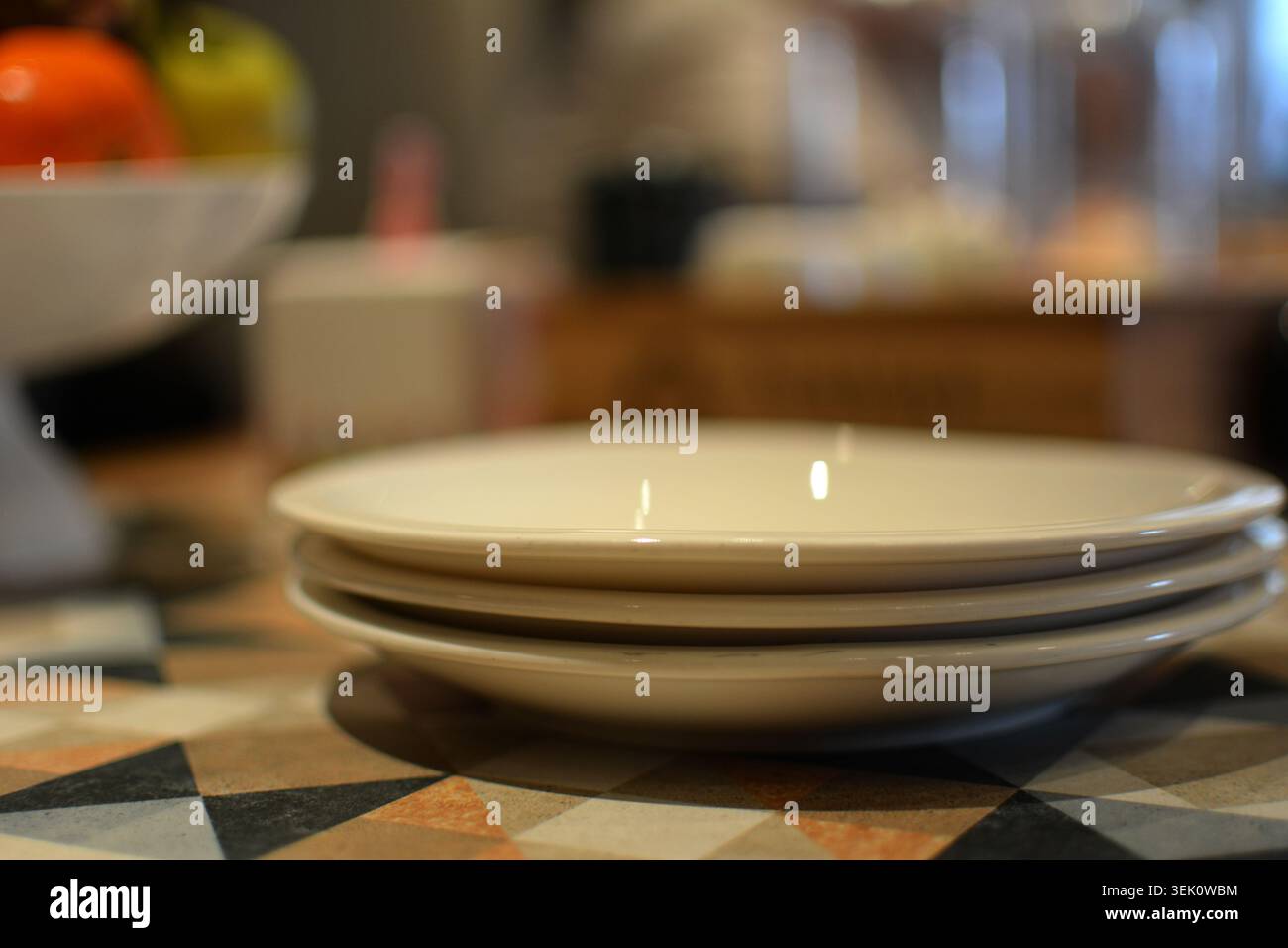 Close-up of three stacked white ceramic plates on a decorative surface with blurred showing colorful fruit and kitchen items. Stock Photo