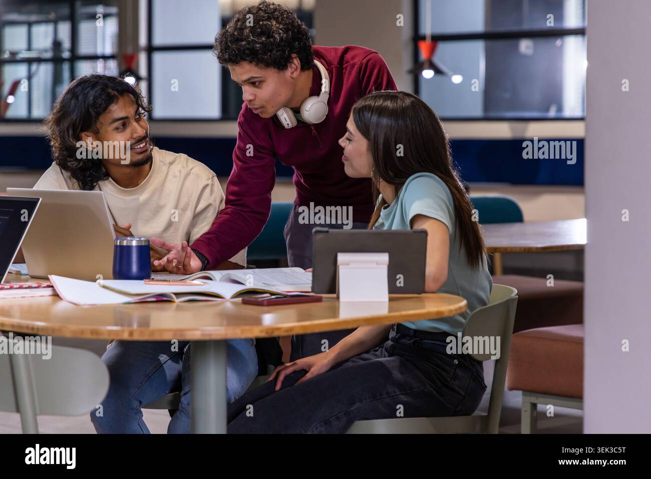 Diverse students collaborating in study lounge around round table with laptop and textbook Stock Photo