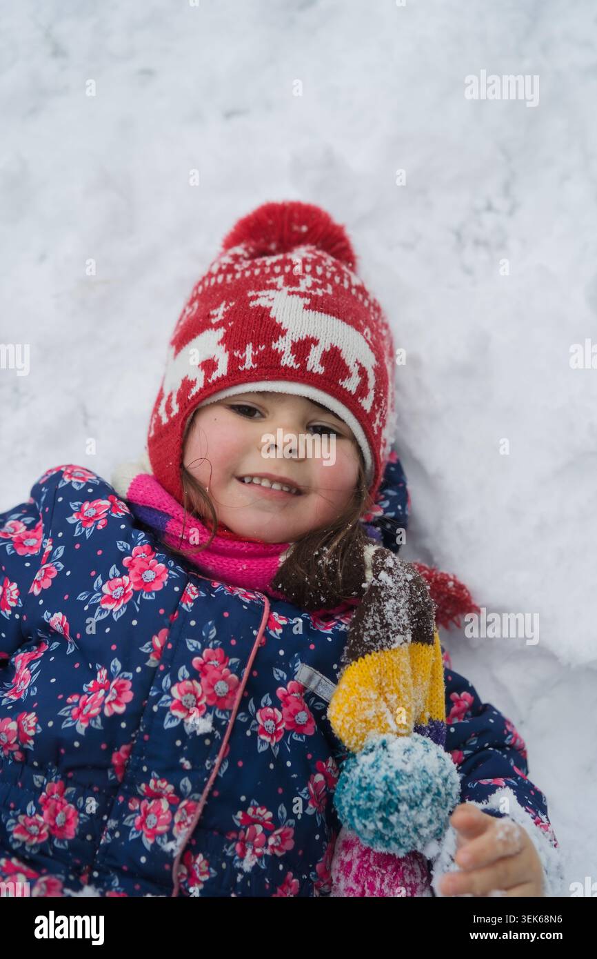 Portrait of smiling girl in red hat lying in snow holding mitten Stock Photo