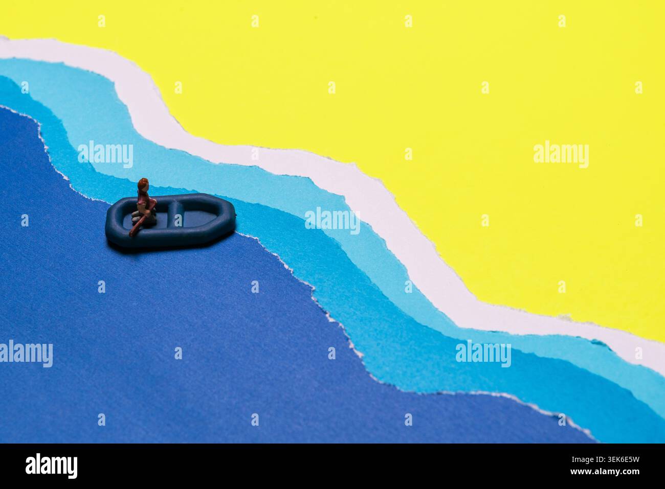 Miniature Person Seated In Small Dinghy Boat Floating On Blue Wavy Water Against Yellow Sky Backdrop Stock Photo