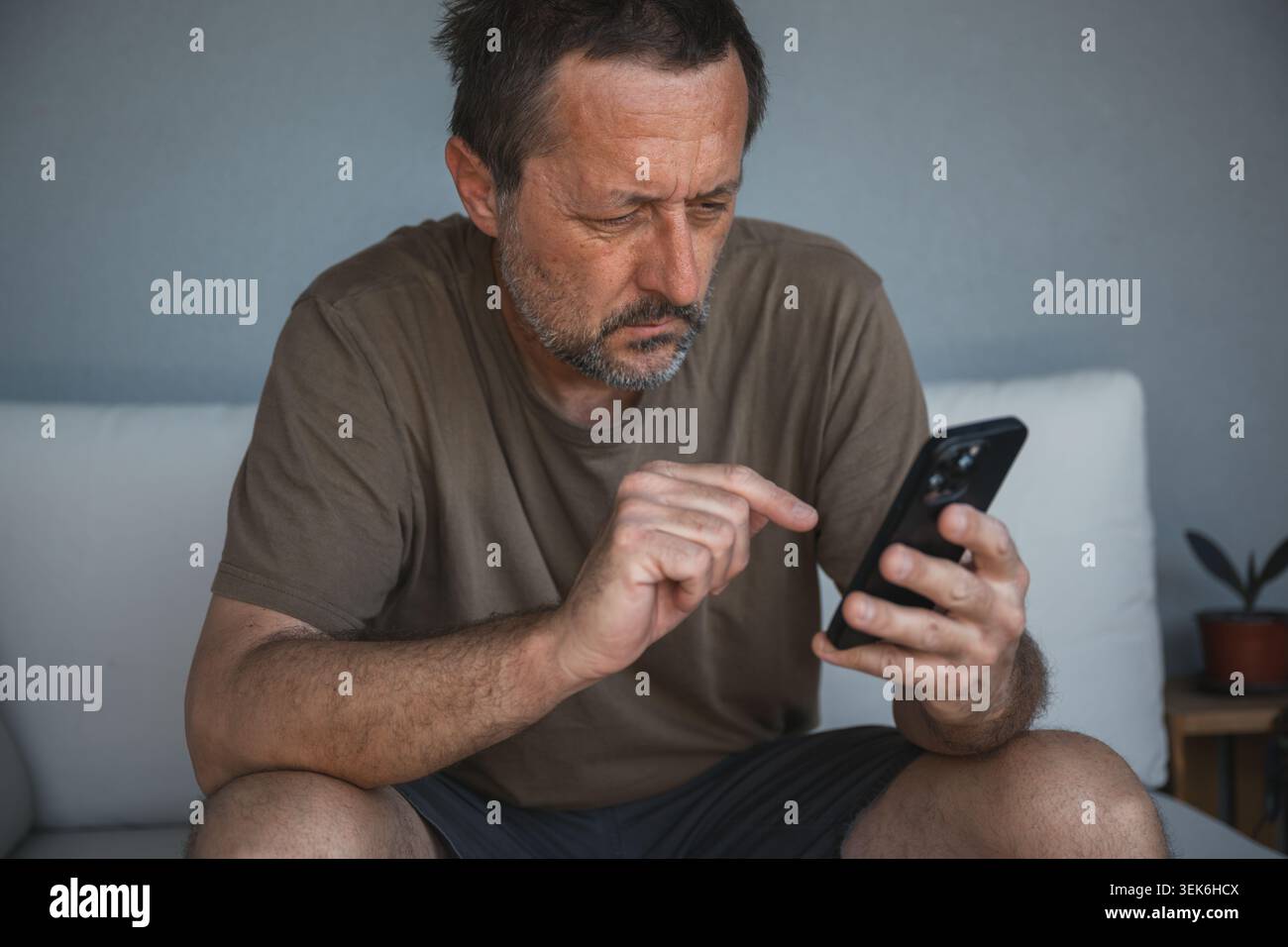 Focused middle-aged man using smartphone on couch at home checking messages and browsing, selective focus Stock Photo