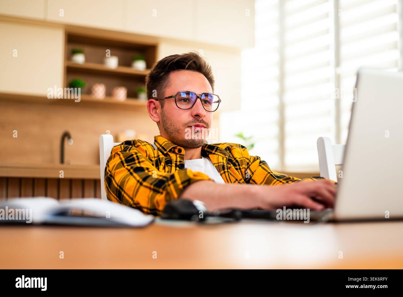 With a pensive expression, a man types on his laptop, appearing to analyze information or solve a problem online. Stock Photo