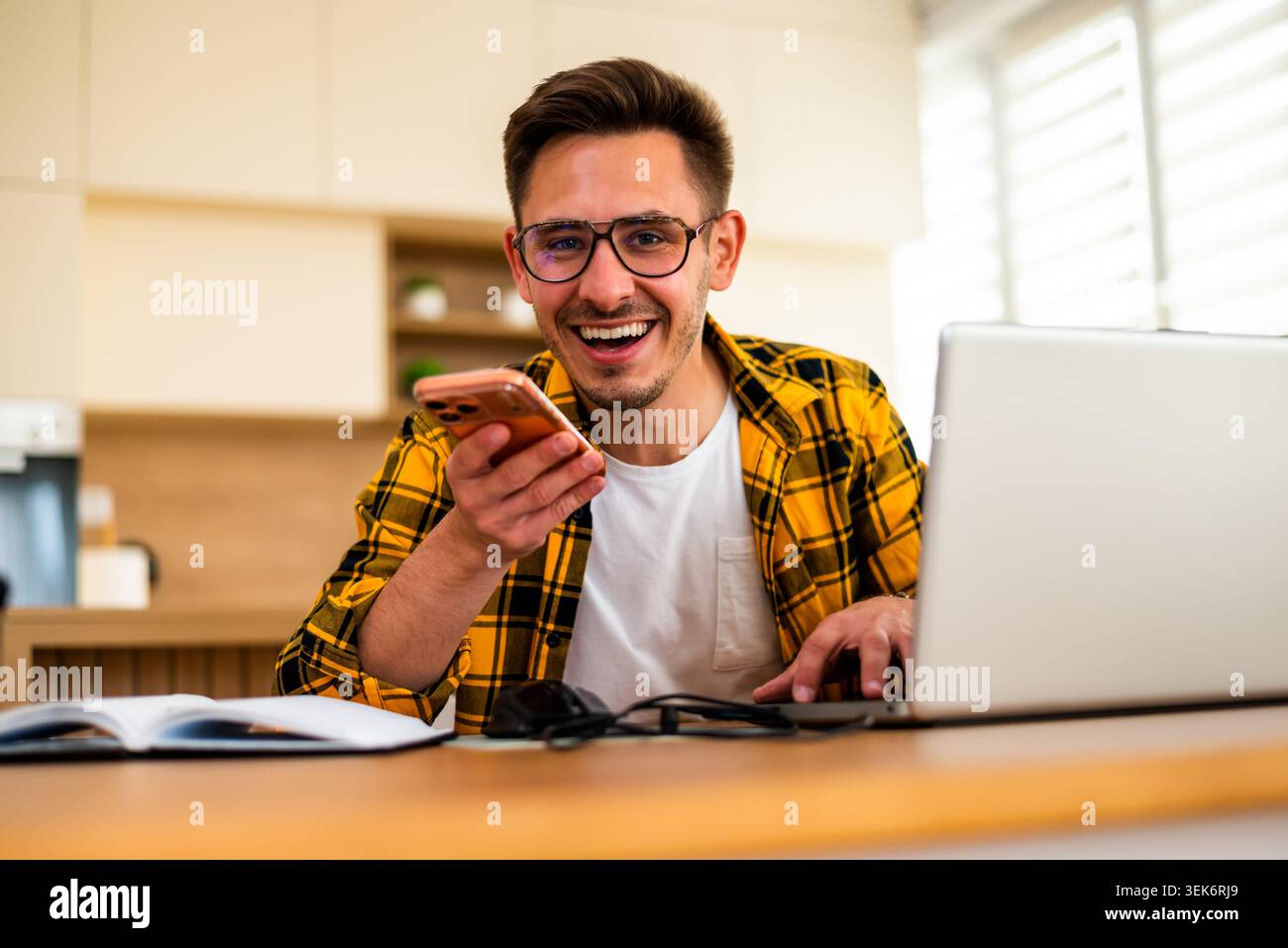 Happy young man is working from his home. He is using laptop and talking on smartphone. Stock Photo