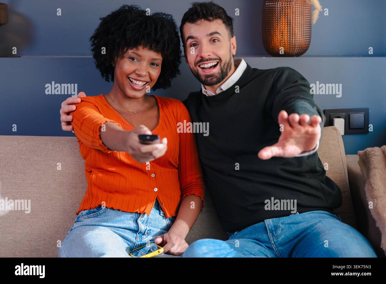 Happy multiracial couple watching television and using remote control on sofa Stock Photo